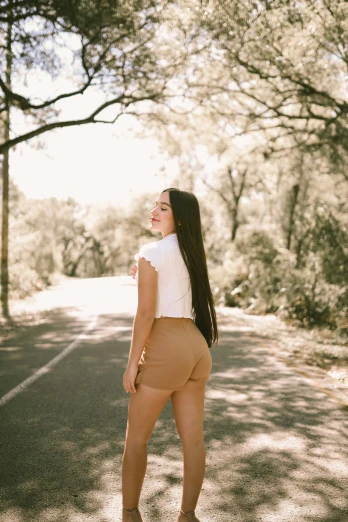 a woman standing in the middle of a road, by Robbie Trevino, tan shorts, brown colored long hair, against the backdrop of trees, facing away