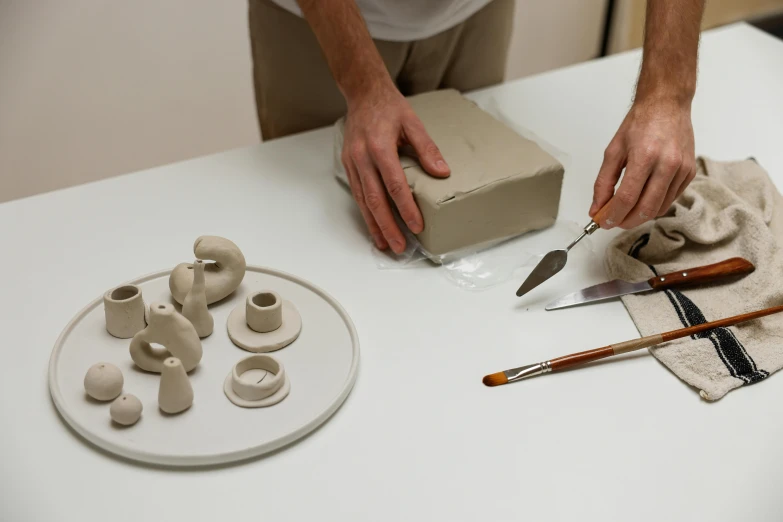 a man is making clay objects on a table, a still life, inspired by Hendrik Gerritsz Pot, porcelain holly herndon statue, multi-part, grey, product introduction photo