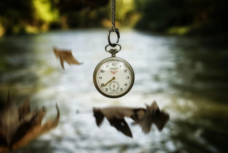 a close up of a clock hanging from a chain, an album cover, inspired by Pierre Pellegrini, trending on pexels, happening, on a riverbank, falling leaves, pocket watch, view