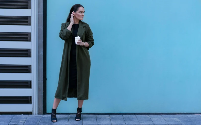 a woman standing in front of a blue wall talking on a cell phone, inspired by Marina Abramović, pexels contest winner, trench coat, she wears a dark green dress, manuka, army girl outfit