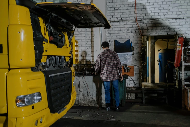 a man standing next to a yellow truck in a garage, pexels contest winner, arbeitsrat für kunst, man sitting facing away, robot repair workshop, 🚿🗝📝, panorama shot