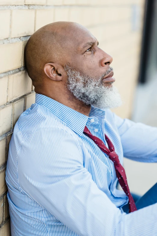 a bald man sitting against a brick wall, a photo, inspired by George Barret, Sr., trending on pexels, grey trimmed beard, contemplation, wearing a shirt with a tie, african man