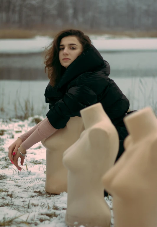 a woman sitting next to a row of mannequins in the snow, an album cover, inspired by Elsa Bleda, pexels contest winner, still from a music video, portrait sophie mudd, ceramics, near a lake