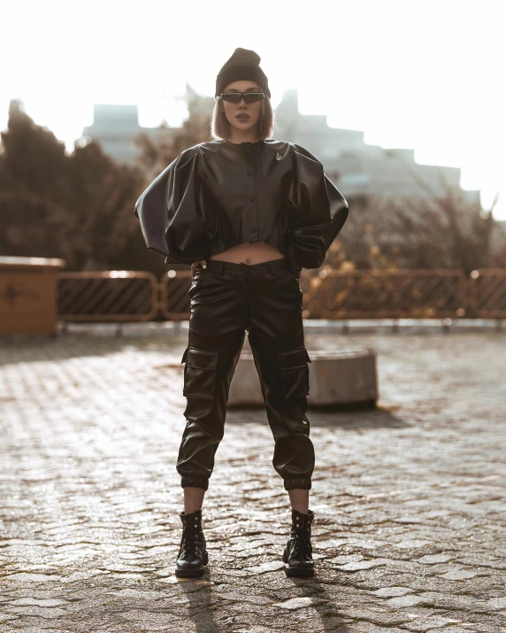a woman standing in the middle of a brick road, an album cover, by Emma Andijewska, trending on pexels, graffiti, leather pilots uniform, baggy pants, techwear clothes, black leather costume