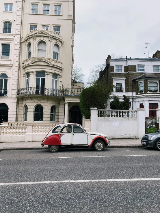 a red and white car driving down a street next to a tall building, a photo, pexels contest winner, neoclassicism, white houses, 😭 🤮 💕 🎀, london, snail