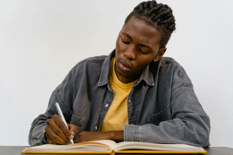 a man sitting at a table writing on a book, a drawing, pexels contest winner, black teenage boy, over the shoulder, plain background, background image