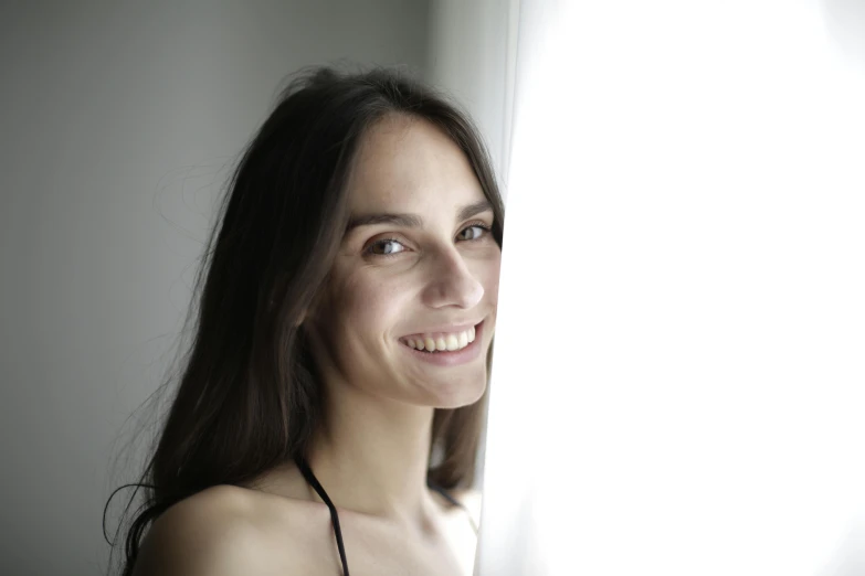 a woman standing in front of a window smiling, pexels contest winner, antipodeans, girl with dark brown hair, close - up studio photo, very thin, looking up at camera