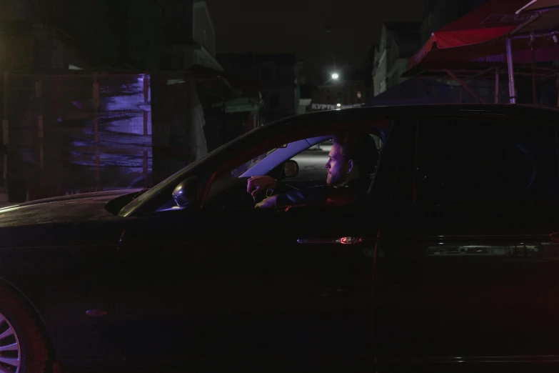 a man sitting in the driver's seat of a car, overlooking a dark street, ariel perez, aura jared and wires, lights on