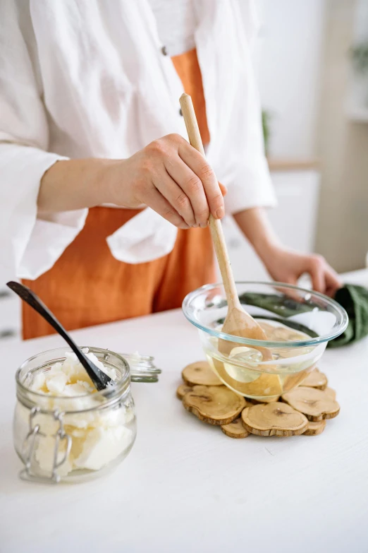 a woman in an orange apron mixing ingredients in a bowl, inspired by Li Di, trending on pexels, butter sculpture, glass jar, square, white