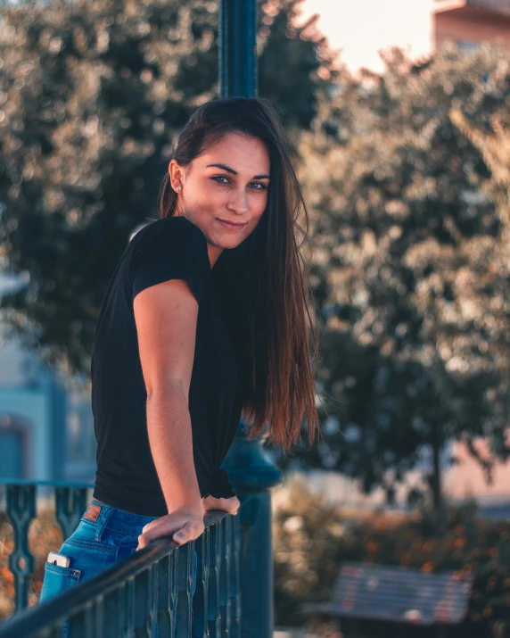 a woman leaning on a railing with trees in the background, pexels contest winner, happening, she is wearing a black tank top, attractive girl tomboy, fair olive skin, grinning lasciviously