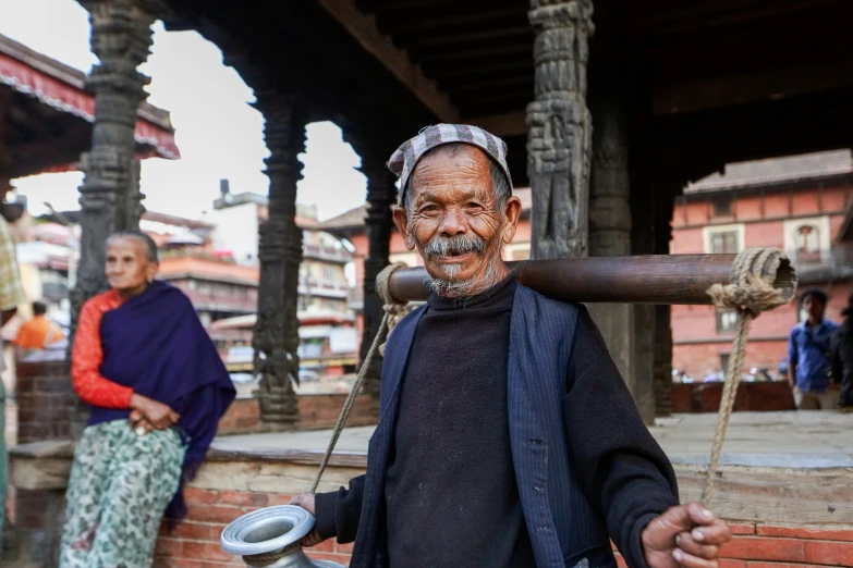 a man standing in front of a building with a stick on his shoulder, nepali architecture buildings, two old people, liam brazier, holding a thick staff