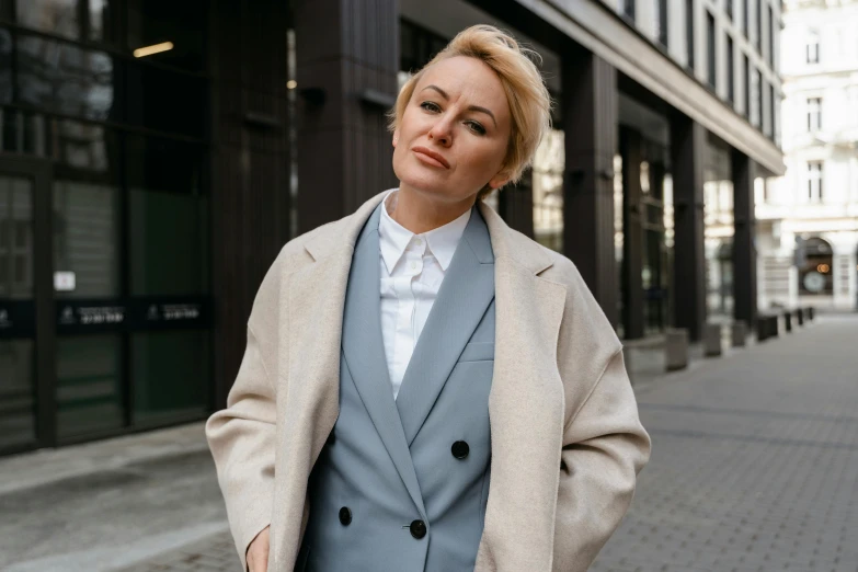 a woman standing on a sidewalk in front of a building, trending on pexels, trench coat and suit, blonde swedish woman, blue and gray colors, thumbnail