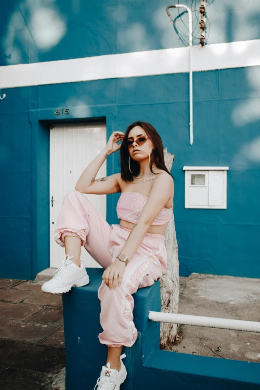 a woman sitting on a bench in front of a blue building, an album cover, inspired by Elsa Bleda, trending on pexels, pink clothes, croptop, with sunglass, wearing a track suit