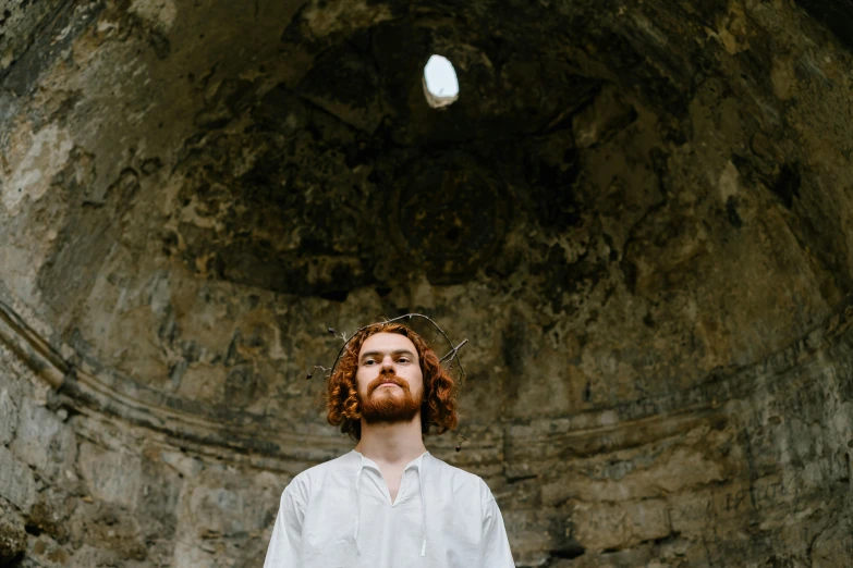 a man standing in front of a stone wall, an album cover, by Attila Meszlenyi, pexels contest winner, renaissance, ginger hair, holy ceremony, white stone arches, curls on top of his head