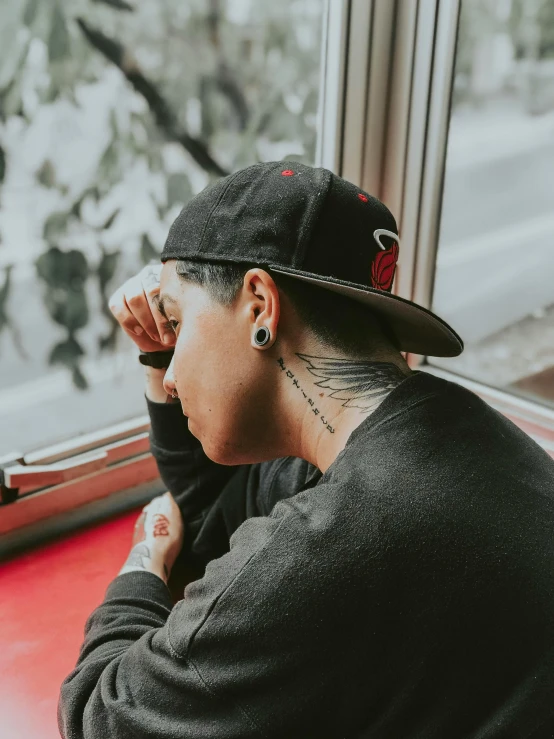 a man sitting at a table looking out a window, a tattoo, inspired by Seb McKinnon, pexels contest winner, he has a red hat, boy has short black hair, profile image, woman in streetwear