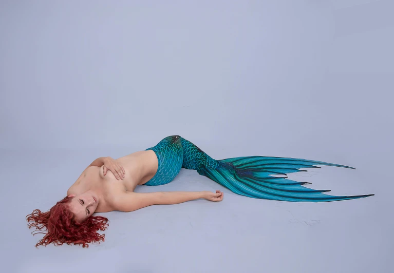a woman laying on the ground with a mermaid tail, inspired by Evelyn De Morgan, hurufiyya, lycra costume, on a pale background, teal, multicoloured