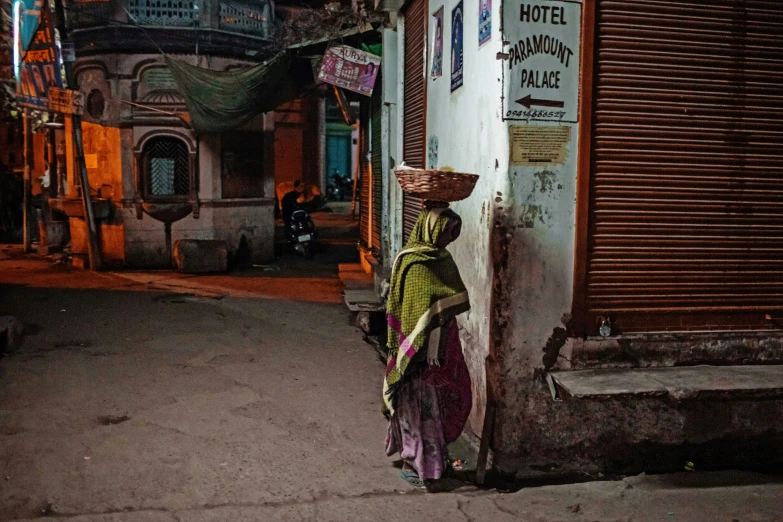 a woman walking down a street with a basket on her head, a picture, inspired by Steve McCurry, pexels contest winner, night color, india, old shops, instagram post