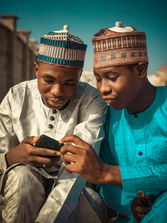 two men sitting next to each other looking at a cell phone, trending on pexels, somali attire, a boy, 2030, multicoloured