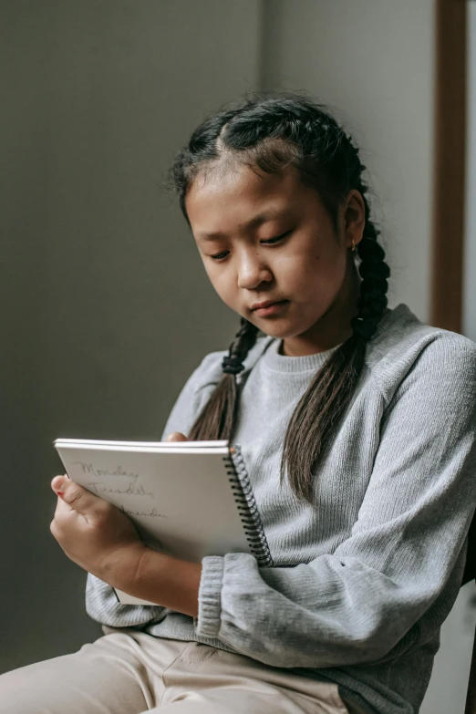 a young girl sitting on a chair using a tablet, inspired by Ruth Jên, pexels contest winner, holding notebook, pensive expression, grey, young asian girl