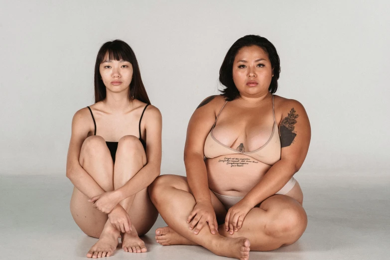 a couple of women sitting next to each other, inspired by Vanessa Beecroft, trending on pexels, her belly is fat and round, half asian, tattooed body, tan skin)