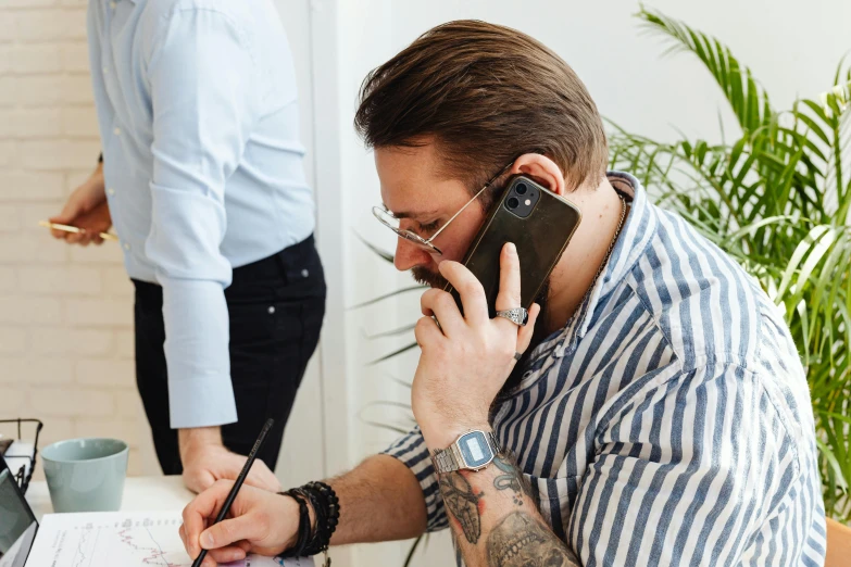 a man sitting at a desk talking on a cell phone, by Adam Marczyński, trending on pexels, arbeitsrat für kunst, tattoo artist, 9 9 designs, writing on a clipboard, taken on iphone 1 3 pro