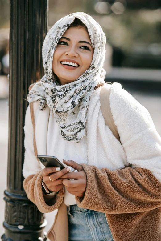 a woman smiles while looking at her cell phone, inspired by Maryam Hashemi, trending on pexels, hurufiyya, wearing a scarf, malaysian, light tan, grey