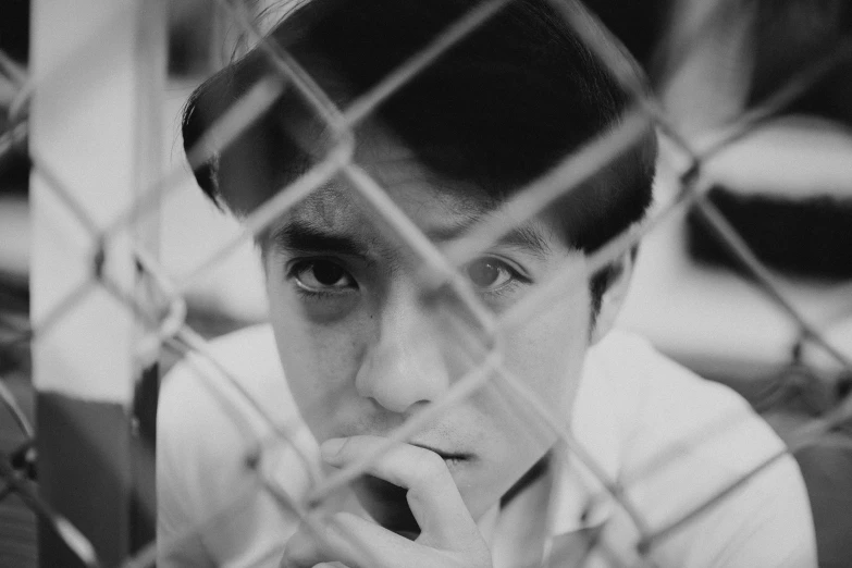 a black and white photo of a boy behind a fence, by Maurycy Gottlieb, in style of monkeybone, ray william johnson, asian man, thoughtful )