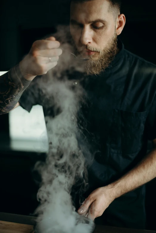 a man standing in a kitchen preparing food, a tattoo, by Austin English, pexels contest winner, swirling wispy smoke, with a beard and a black shirt, standing with a black background, aussie baristas