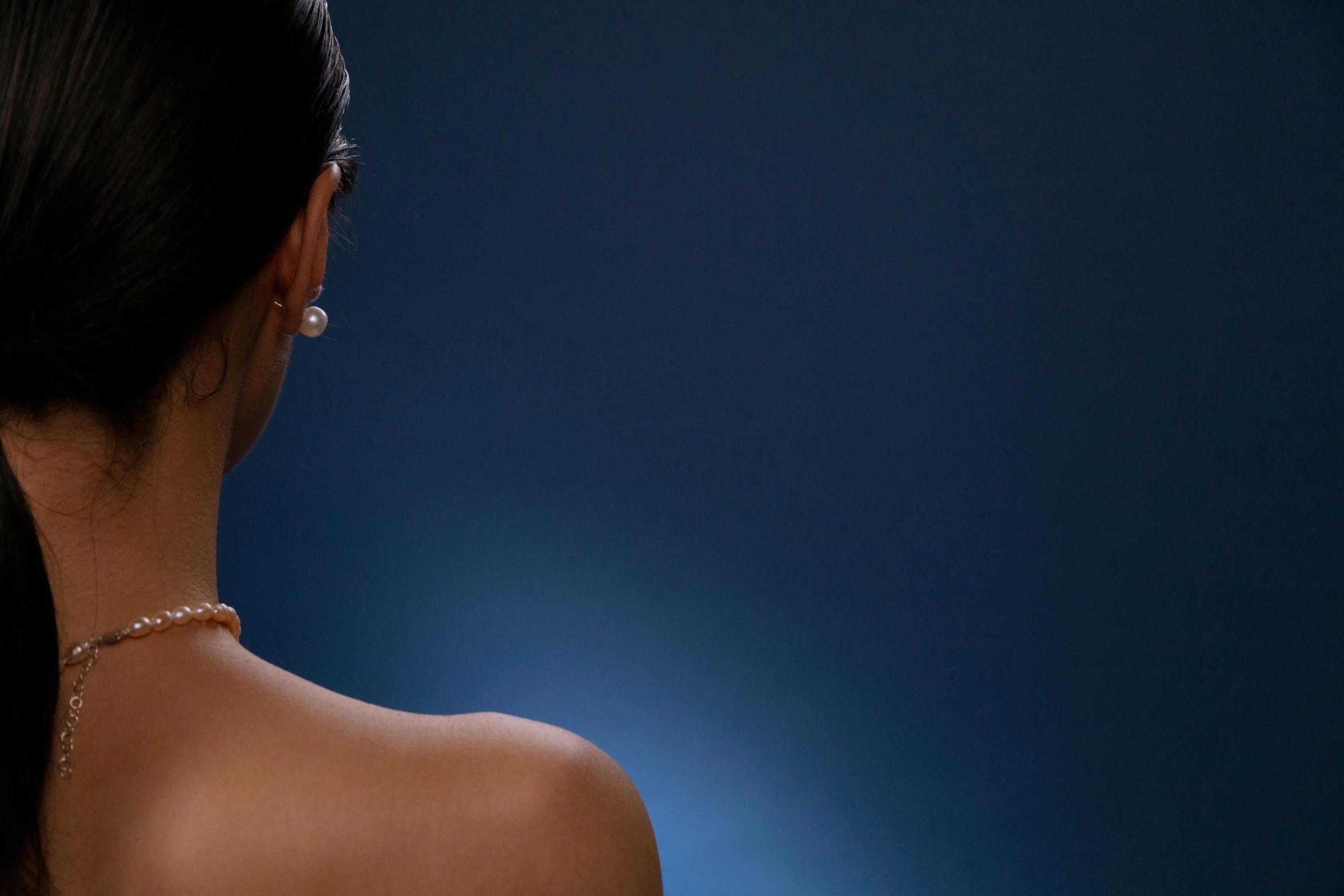 a woman with her back to the camera, soft blue lighting, photoshoot for skincare brand, pearls, deep colour