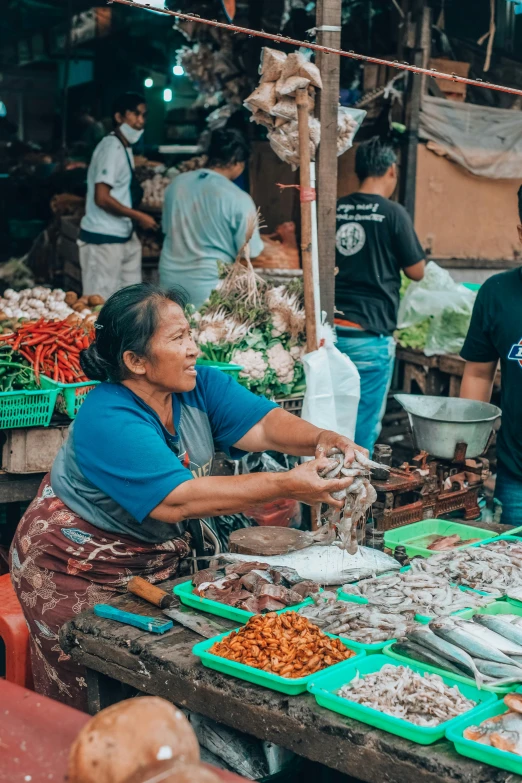 a woman standing in front of a table filled with food, pexels contest winner, sumatraism, busy market, two hands reaching for a fish, square, inspect in inventory image