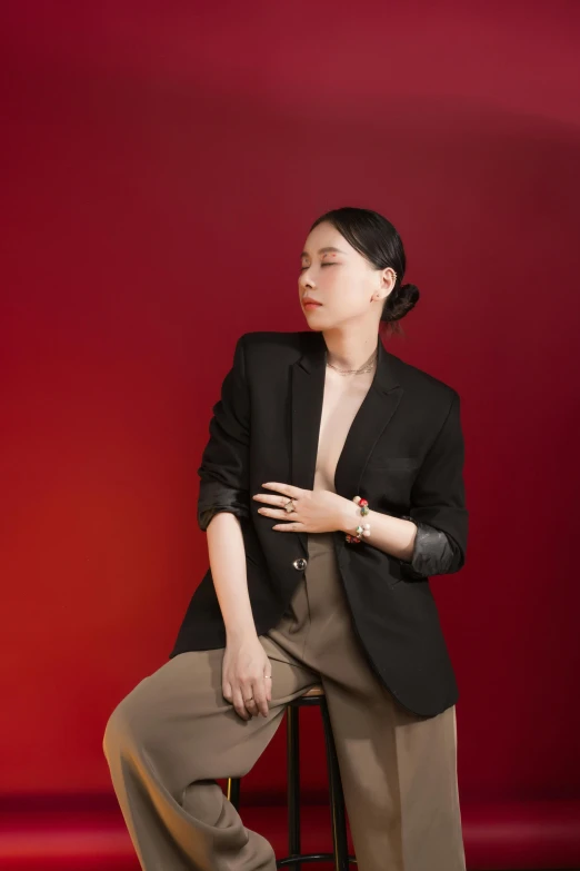 a woman sitting on a stool in front of a red wall, an album cover, inspired by Ruth Jên, trending on pexels, subject detail: wearing a suit, jingna zhang, androgynous person, jewelry