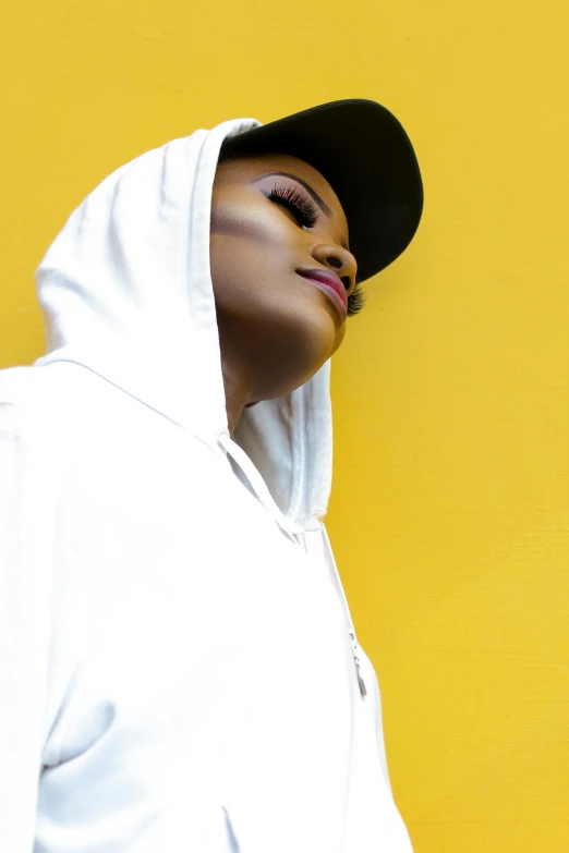 a woman standing in front of a yellow wall, an album cover, by Chinwe Chukwuogo-Roy, pexels, wearing white silk hood, hat and hoodie, profile posing, dazzling lights