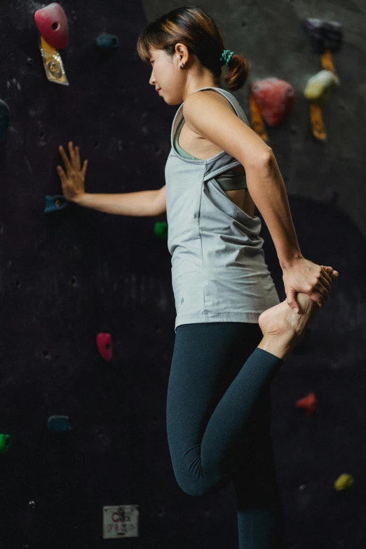 a woman standing in front of a climbing wall, arabesque, wearing fitness gear, grey, full frame image, leggings