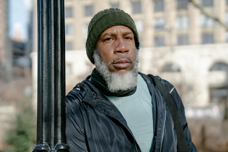 a close up of a person holding a pole with a building in the background, an album cover, inspired by Gordon Parks, pexels contest winner, realism, he also wears a grey beanie, silver full beard, harlem, looking directly at the camera