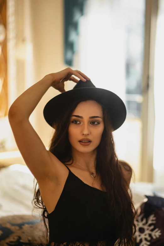 a woman sitting on top of a bed wearing a hat, a picture, trending on pexels, wearing black camisole outfit, young middle eastern woman, tall forehead, thick jawline