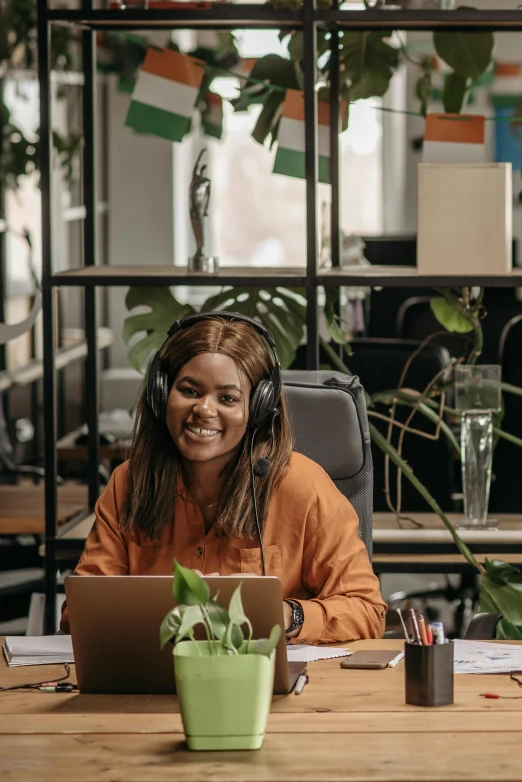 a woman sitting at a table with a laptop, wearing black headphones, kano), in an office, smiling playfully