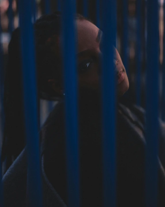 a woman standing in front of a blue fence, inspired by Elsa Bleda, trending on unsplash, happening, sitting in a dark prison cell, dark skin, low quality footage, lgbtq