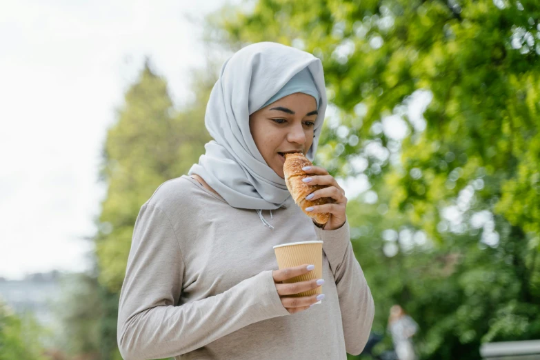 a woman in a hijab eating a donut and drinking coffee, inspired by Modest Urgell, shutterstock, hurufiyya, people on a picnic, holding toasted brioche bun, australian, 2019 trending photo