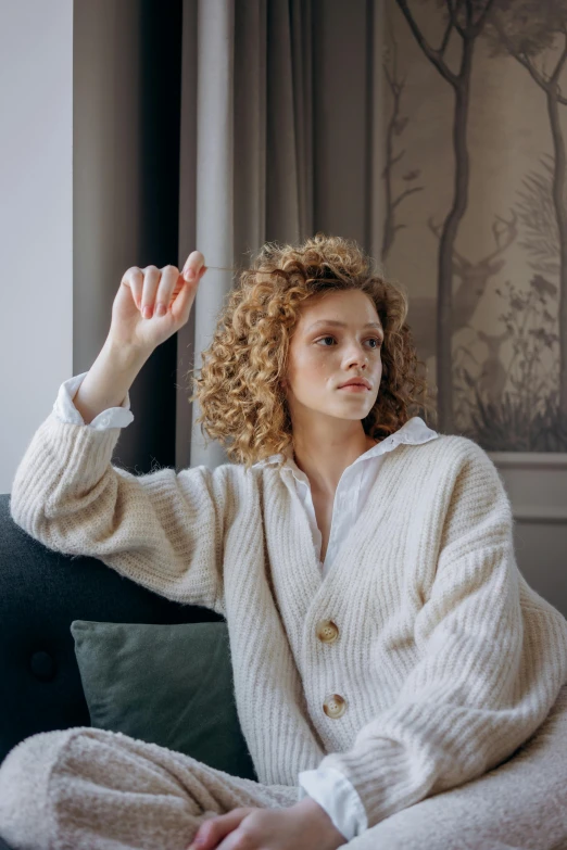 a woman sitting on top of a couch next to a window, by Anna Boch, trending on pexels, curly middle part haircut, cardigan, waving, non binary model