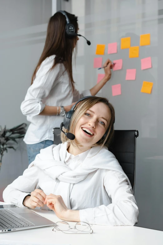 a woman sitting at a desk in front of a laptop, trending on pexels, arbeitsrat für kunst, earing a shirt laughing, whiteboards, wearing headset, woman holding another woman