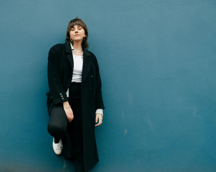a woman standing in front of a blue wall, an album cover, by Winona Nelson, pexels contest winner, suprematism, wearing black overcoat, brown hair and bangs, white and black clothing, leaning on the wall