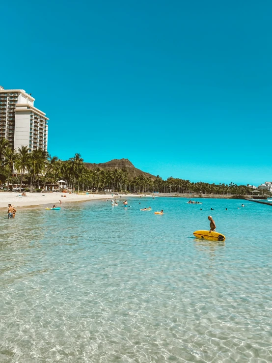 a man riding a surfboard on top of a sandy beach, posing in waikiki, crystal clear blue water, people swimming, corn floating in ocean