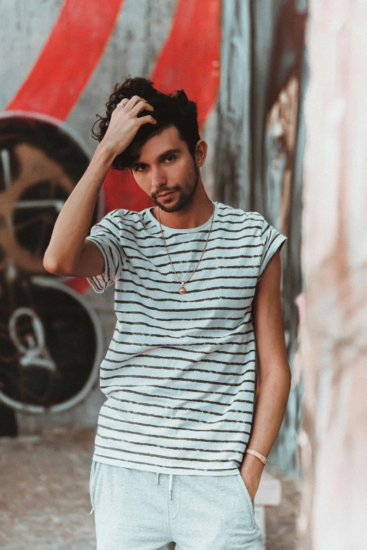 a man standing in front of a graffiti covered wall, an album cover, inspired by Germán Londoño, trending on pexels, striped shirt, beautiful young man, wearing a t-shirt, androgyny