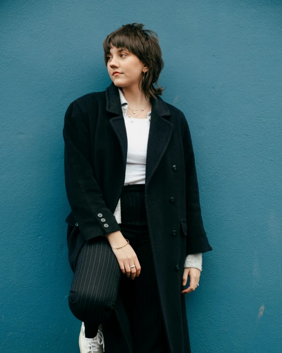 a woman standing in front of a blue wall, an album cover, inspired by Elsa Bleda, trending on pexels, suprematism, wearing black overcoat, portrait androgynous girl, pinstripe suit, ( ( wearing a long coat ) )