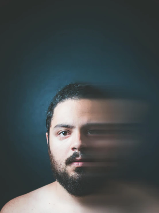 a man with a beard standing in front of a dark background, by Alejandro Obregón, with haunted eyes and dark hair, pareidolia, lgbtq, blurry background