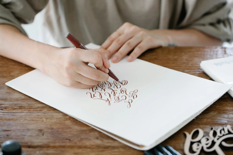 a person sitting at a table writing on a piece of paper, a drawing, inspired by Ruth Jên, trending on pexels, letterism, decorative art, brown, calligraphy, uppercase letter