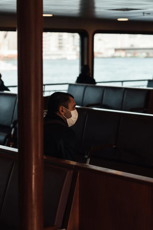 a person wearing a face mask on a boat, interior of staten island ferry, man sitting facing away, light and dark, brown