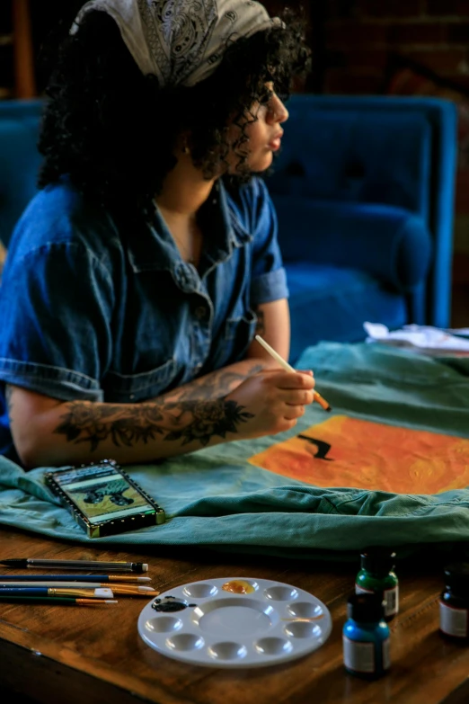 a woman sitting at a table working on a painting, a detailed painting, trending on pexels, artist wearing overalls, marijuana organic painting, teal and orange colours, painting on a badge