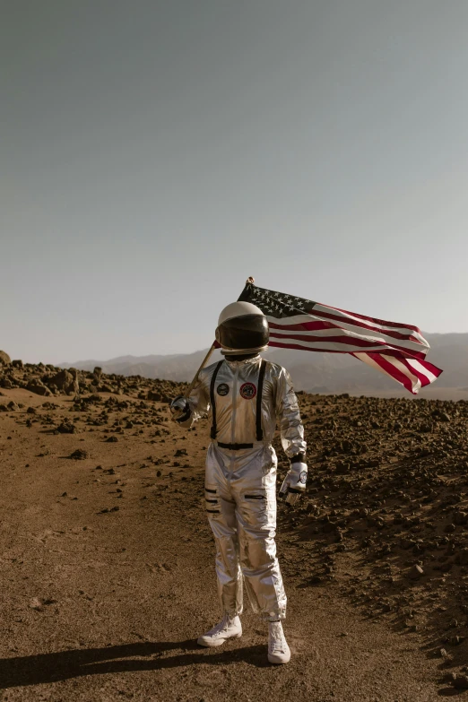 a man standing on a dirt road holding an american flag, inspired by Alan Bean, unsplash contest winner, on mars, movie still 8 k, red sea, woman astronaut