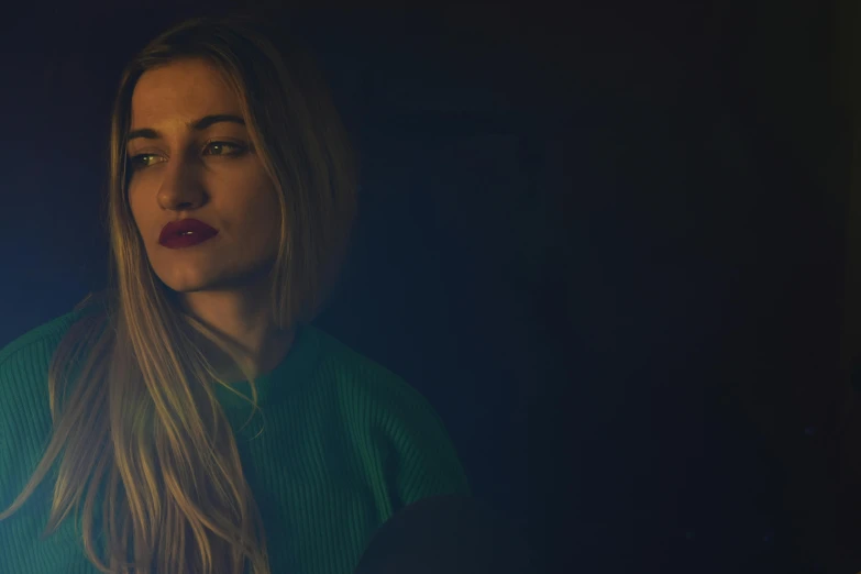 a woman standing in front of a blue background, a portrait, inspired by Elsa Bleda, pexels, realism, very dark with green lights, blonde women, concert photo, atmospheric warm colorgrade